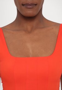 Gros plan d'une personne portant un top sans manches orange vif avec un décolleté carré, montrant les épaules et le haut de la poitrine.