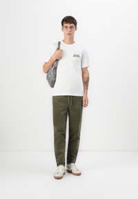 T-shirt bianco in cotone con logo, pantaloni in corduroy verde oliva, borsa grigia e sneakers bianche con dettagli marroni. Outfit semplice e casual.