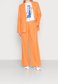Oranžový oblek s přiléhavým sako a širokými kalhotami, doplněný bílým tričkem s grafikou a bílými sandály na vysokém podpatku. Hladká textura.