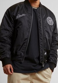 La veste bomber noire se distingue par ses poignets côtelés, sa fermeture éclair et ses logos brodés. Confectionnée dans un tissu lisse avec une texture légèrement brillante.