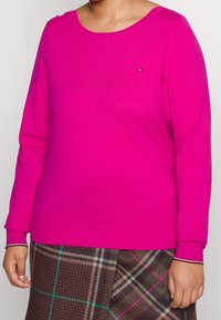Suéter fucsia de manga larga y cuello redondo. Presenta un pequeño logotipo y puños acanalados. Combinado con una falda texturizada de cuadros.