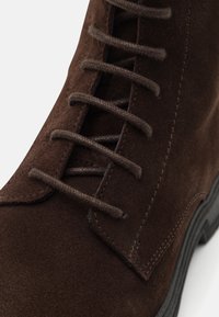 Botín de ante marrón con cordones, con costuras detalladas y cordones redondos, inclinado para mostrar la parte superior y parte de la suela negra.