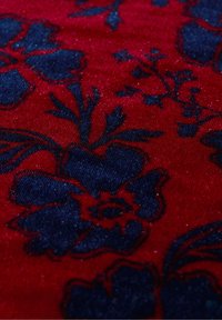 Röd tyg med ett mörkblått blommönster och intrikata detaljer. Texturen verkar mjuk med en subtil glans.