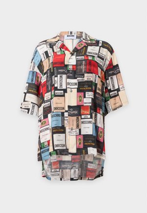 Das Modell trägt ein locker sitzendes, passendes Hemd und eine Hose mit bunten Moschino-Logo-Aufdrucken auf weißem Hintergrund.