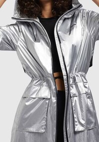 Srebrna wodoodporna kurtka z kapturem, ściąganym w pasie, z zamkiem błyskawicznym z przodu i dwiema dużymi kieszeniami, o błyszczącej, gładkiej teksturze.