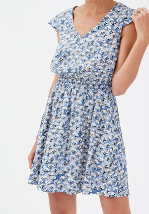 Femme portant une robe bleu sans manches avec une taille élastique et un motif floral blanc et noir en éventail, posant avec une main près de l'épaule.
