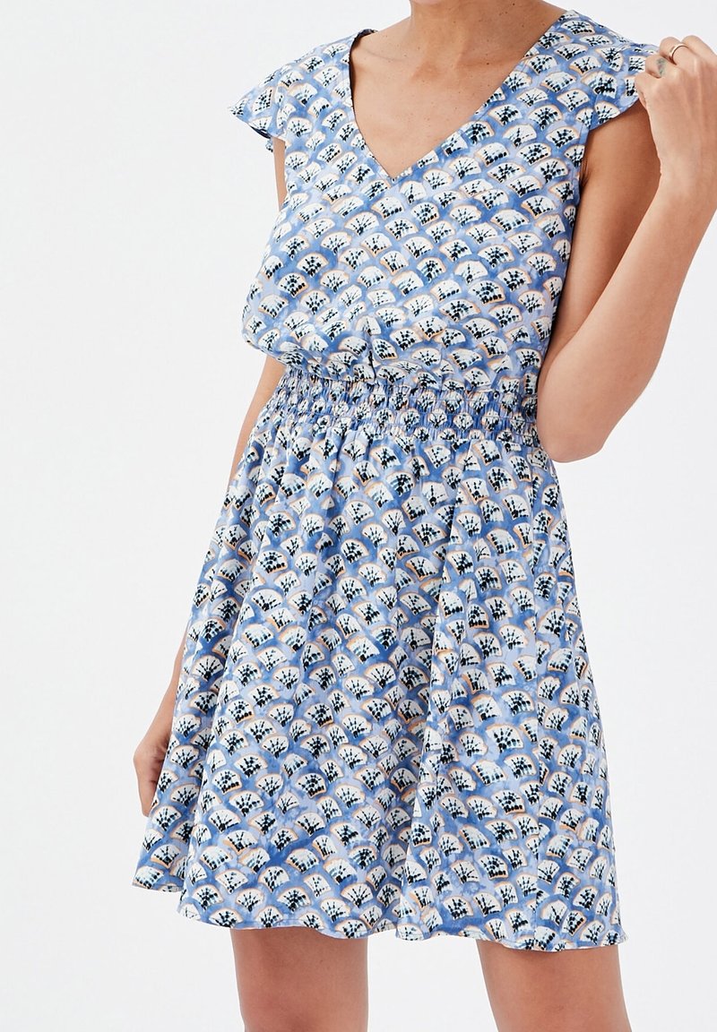 Femme portant une robe bleu sans manches avec une taille élastique et un motif floral blanc et noir en éventail, posant avec une main près de l'épaule.