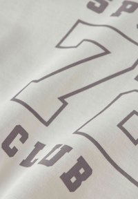 Vitt tyg med stort konturerat nummer "72" och mindre texten "CLUB" tryckt i mörkgrått. Tygstruktur och svaga veck syns.