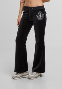 Czarne dresowe spodnie z weluru z elastycznym paskiem, rozszerzanymi nogawkami i białym haftowanym logo na przedniej kieszeni. Miękka tekstura.