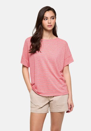 Joven mujer con el pelo largo y oscuro, vestida con una camiseta de manga corta a rayas rojas y blancas y pantalones cortos beige, de pie con una mano en el bolsillo.