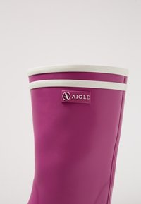 Rosa gummistövlar med vita horisontella ränder upptill och en logotags. Ytan är slät med en rundad övre kant.