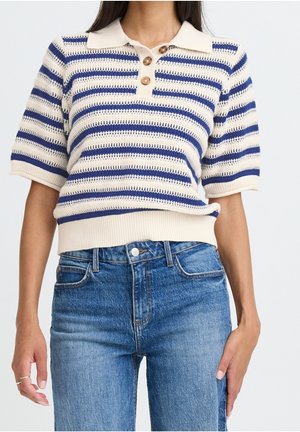 Femme portant un haut tricoté à manches courtes bleu et blanc à rayures avec des boutons en bois, associé à un jean en denim bleu taille haute.