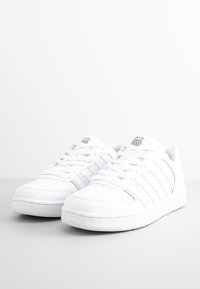 Bílé sportovní boty s hladkým koženým povrchem, šesti dírami na tkaničky a jemnými šedými akcenty na pruzích. Plochá gumová podrážka.
