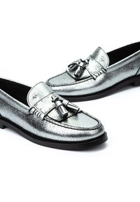 Mocasines metálicos plateados con un acabado texturizado, que presentan un acento decorativo de borlas y detalles de costura en una suela de goma negra.