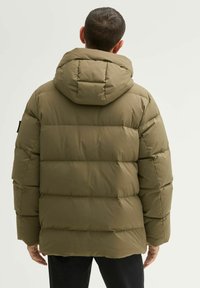 Oliwkowa pikowana kurtka z kapturem, z poziomymi przeszyciami, gładką fakturą i detalem naszywki na lewym rękawie.