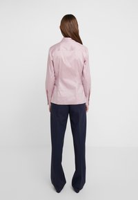 Camicia rosa chiaro con colletto, maniche lunghe e design aderente, abbinata a pantaloni larghi color blu scuro. Tessuto liscio, dettagli minimali.