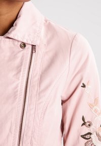 Světle růžová bomber bunda s stojáčkem, viditelným kovovým zipem a květinovou výšivkou na rukávu. Hladká textilie.