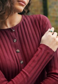 Top bordeaux a coste con scollo rotondo, cinque bottoni scuri, maniche lunghe e dettagli in tessuto strutturato. Due anelli visibili sulla mano.