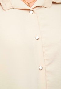 Beige skjorte med knapper som har en glatt tekstur, klassisk krage og sølvknapper langs fronten.