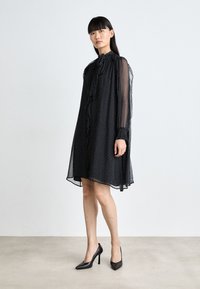 Schwarzes Pünktchenkleid aus transparenter Stoff, mit hohem Kragen, Rüschendetails und langen Ärmeln, kombiniert mit schwarzen hohen Schuhen.