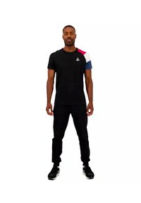 Zwart sportt-shirt met kleurgeblokkeerde schouders in rood, wit en blauw. Gepaard met zwarte broek en zwarte sneakers.