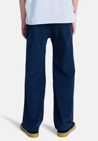 Osoba ubrana w luźne, ciemnoniebieskie spodnie sztruksowe i czarne trampki z gumowymi podeszwami, stojąca na jednolitym białym tle.