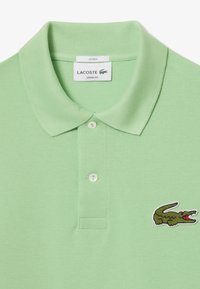 Ljusgrön polo med texturerad tyg, tre-knappsknäppning och broderad krokodil-logga på vänster bröst. Unisexdesign.