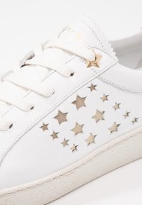 Vita sneakers med vita snören, stjärnformade utskärningar på sidan och en guldstjärnformad ögjure nära toppen.