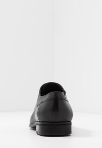 Chaussure en cuir noire avec une finition lisse, de forme arrondie, à talon bas et avec des accents texturés au niveau de la cheville. Design simple et classique.