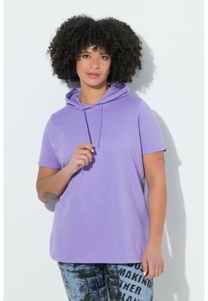 Person mit lockigem Haar, die einen kurzärmligen lavendelfarbenen Hoodie und blaue gemusterte Hosen mit fettem schwarzem Text trägt, steht vor weißem Hintergrund.