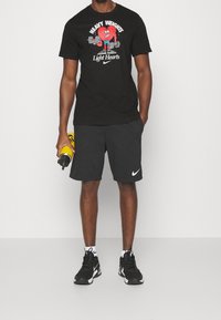 Czarny t-shirt z grafiką, na którym znajduje się czerwony rysunkowy bohater z ciężarami, zestawiony z czarnymi szortami Nike i czarnymi butami sportowymi, trzymający żółtą butelkę na wodę.