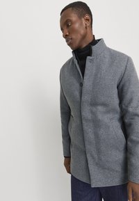 Un homme portant un manteau gris à boutons sur un col roulé noir et un pantalon sombre, se tenant contre un fond clair et uni.