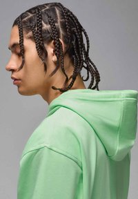 Felpa verde con una finitura testurizzata, dotata di cappuccio con coulisse e dettagli di cucitura visibili, indossata da una persona con capelli trecciati.