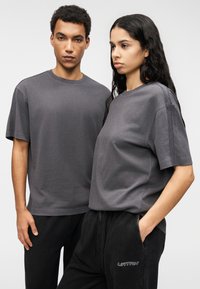 Γκρι βαμβακερά t-shirt με άνετη εφαρμογή και κοντά μανίκια, με διακριτικές ραφές, φορεμένα με μαύρα φόρμας παντελόνια.