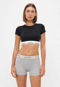 Crni pamučni crop top s kratkim rukavima i bijelim Calvin Klein logom; sivi pamučni bokser šorts s bež pojasom na kojem je logo.