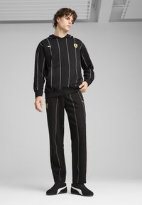 Crna dukserica s bijelim vertikalnim prugama i malim Ferrari logom, u kombinaciji s crnim hlačama koje također imaju pruge, sve od mekog materijala.