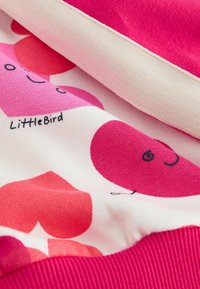 Material colorat cu forme abstracte roz și roșii și o față zâmbitoare. Textură moale cu tiv solid roz cu dungi. Eticheta poartă inscripția "LittleBird".