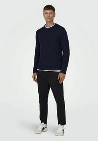 Man staat in een marineblauwe kabelgebreide trui, zwarte broek en witte sneakers met zwarte details, voor een effen witte achtergrond.