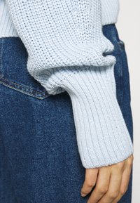 Vaaleansininen neulepuseron hiha, jossa on paksu ribbaus ja teksturoitu kuvio, yhdistettynä tummiin farkkuihin. Lähikuva, joka korostaa yksityiskohtia.