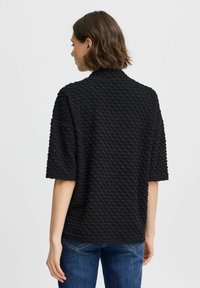 Schwarze Bluse mit Struktur, lässiger Passform, kurzen Ärmeln und einem dezenten Muster. Der Stoff wirkt weich mit einer erhabenen, strukturierten Oberfläche.