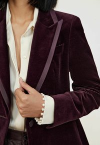 Burgundy bársony blézer selyem részletekkel, egy zsebbel, egy elefántcsont színű gombos ing mellett. Gyöngy karkötő látható a csuklón.