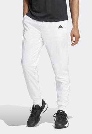 Persona con pantalones deportivos blancos de Adidas, zapatillas negras de Adidas y camiseta negra, de pie sobre un fondo gris claro.