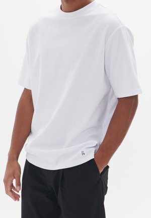 Homme portant une chemise blanche unie à manches courtes et un pantalon noir, avec une main dans la poche et un petit texte imprimé près de l'ourlet.