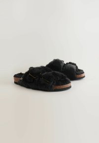 Sorte, fuzzy slippers med en korksål, der har to fremtrædende fluffy remme og diskrete guldfinish på remmene. Blød tekstur overalt.