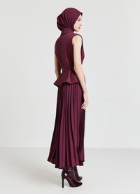 Longue robe bordeaux avec jupe plissée, corsage ajusté et détail peplum. Accompagnée d'un hijab assorti et de bottines à talons.