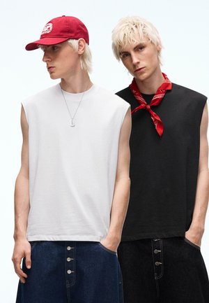 Dwóch młodych mężczyzn o platynowo blond włosach nosi bezrękawniki i spodnie jeansowe; jeden ma czerwoną czapkę i naszyjnik, drugi czerwoną bandanę na szyi.