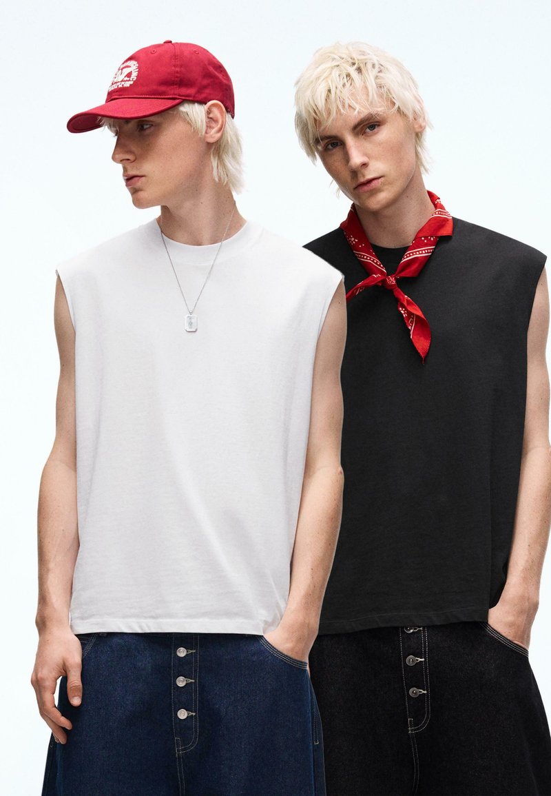 Deux jeunes hommes aux cheveux blond platine portent des débardeurs et des pantalons en denim ; l'un porte une casquette rouge et un collier, l'autre un bandana rouge autour du cou.
