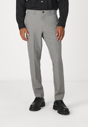 Mężczyzna ubrany w jasnoszare spodnie o wąskim kroju, czarne eleganckie buty i czarną koszulę na guziki, stojący na białym tle.