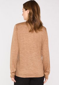 Maglietta a maniche lunghe di colore marrone chiaro con un tessuto texturizzato, orlo arrotondato e polsini aderenti, indossata con pantaloni neri, vista da dietro.