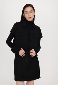 Veste noire ajustée à manches courtes, fermeture à boutons à l'avant et deux poches poitrine, associée à une mini jupe noire texturée.